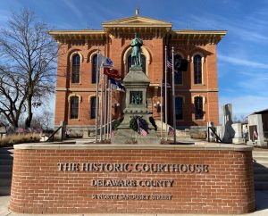 A brick building in the background with a statue of a soldier in the midground and a brick wall in the foreground with the words "The Historic Courthouse - Delaware County - 91 North Sandusky Street" installed on the wall.