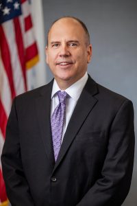 Photo of a man from the waist up looking at the camera. Part of a U.S. flag is visible behind his right shoulder.